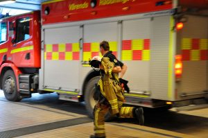 Bild på en brandman som springer mot brandbilen på brandstationen. Han håller i sin skyddsutrustning. Brandbilen syns i bakgrunden. Detta är en genrebild.