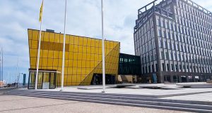 Bild tagen på Linnéuniversitetets bibliotek. Man kan se en liten del av campuset bredvid biblioteket. Biblioteket är gult och framför står det tre stycken flaggstolpar.