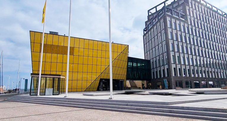 Bild tagen på Linnéuniversitetets bibliotek. Man kan se en liten del av campuset bredvid biblioteket. Biblioteket är gult och framför står det tre stycken flaggstolpar.