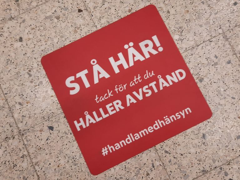 Bild på röd golvdekal inne på ICA MAXI med text: Stå här. Tack för att du håller avstånd.