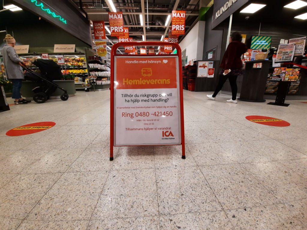 BIld på en skylt inne på ICA MAXI med text: Hemleverans. Tillhör du riskgrupp och vill ha hjälp med handlingen?Vi samarbetar med Svenska Kyrkan som hjälper dig att handla.Ring 0480-421450. Måndag, tisdag, torsdag klockan tio till tolv. Onsdag tretton till femton. Tillsammans hjälper vi varandra! 