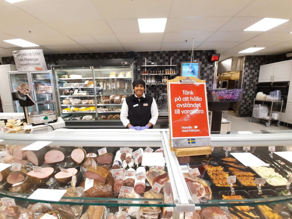 Bild på Veronica Schantz vid delikatessavdelningen. Framför henne står en skylt med text: Tänk på att hålla avståndet till varandra