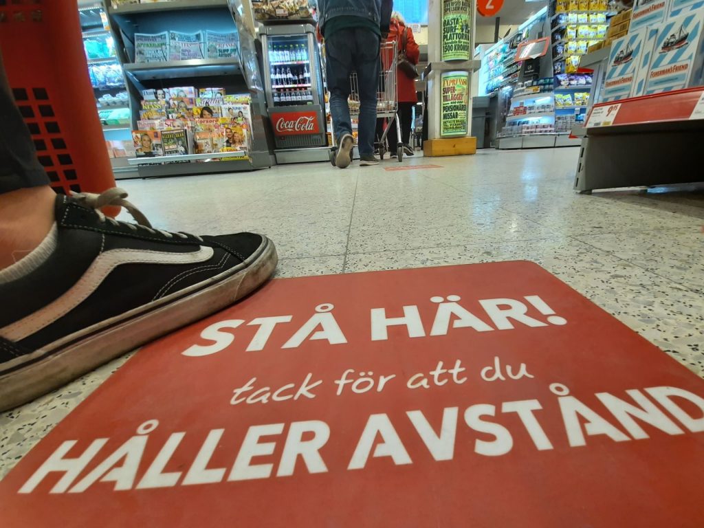 Bild på fot som står på golvdekal på ICA MAXI. På golvdekalen står det: STÅ HÄR! Tack för att du håller avstånd. ett par meter framför ser man två stycken personer längre fram i kön till kassan. 