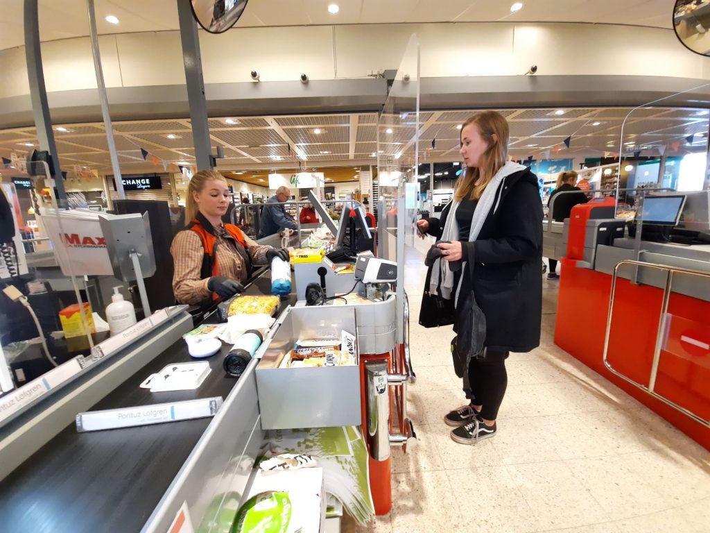 Bild på kassan inne på ICA MAXI. Gabriella Begnert sitter i kassan och Nelly Björkqvist har lagt upp sina varor på bandet. Mellan dem finns ett plexiglas. 
