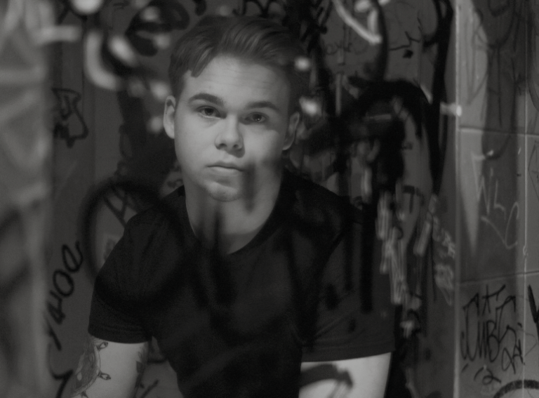 Osscar sitter framför en badrumsspegel som är målat på med svart sprayfärg och kollar in i kameran.