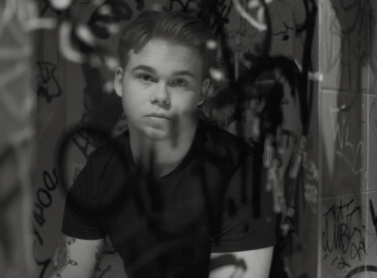 Osscar sitter framför en badrumsspegel som är målat på med svart sprayfärg och kollar in i kameran.