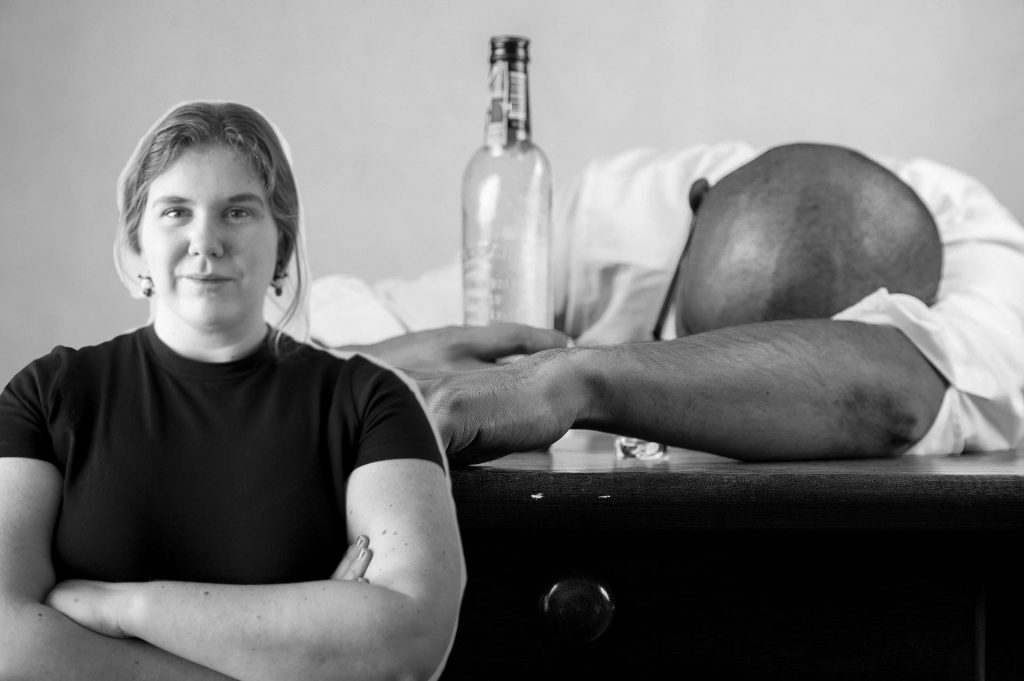 En man sitter med huvudet på bordet hållandes en flaska i andra handen med Sanna Orneklint i förgrunden