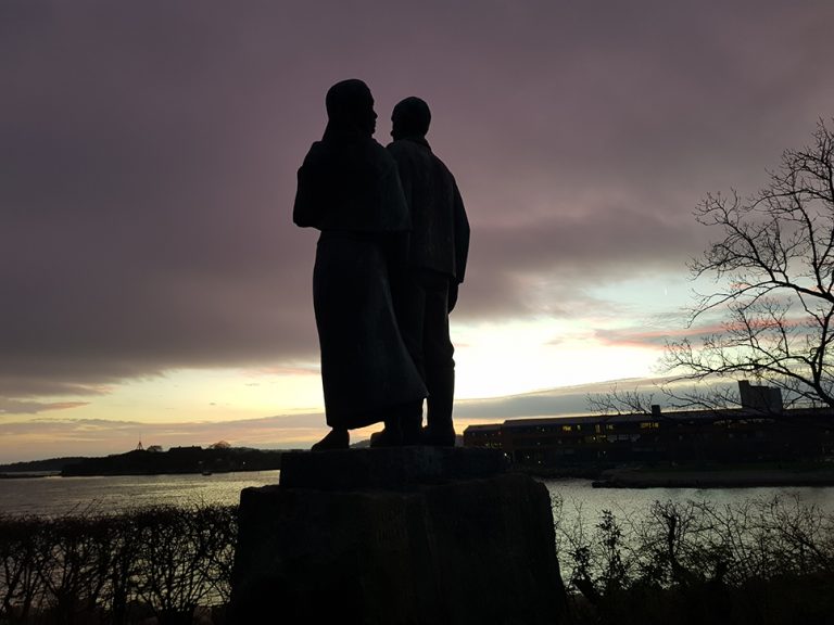 Bild på en staty som föreställer kvinna och en man som går bredvid varandra mer armarna bakom varandras ryggar