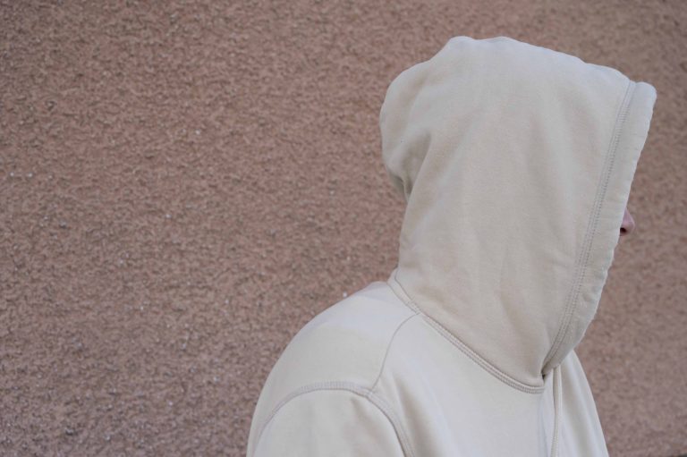 En man står med gul huvtröja mot en rosa husvägg. Han har luvan uppdragen så att man inte ser ansiktet.