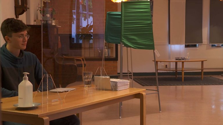 Ett valbås står i mitten av en vallokal. En person sitter vid ett bord bredvid.