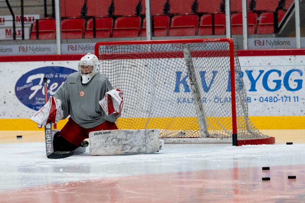 Ishockeymålvakt sittandes framför ishockeymål till vänster i bild. På isen ligger puckar.