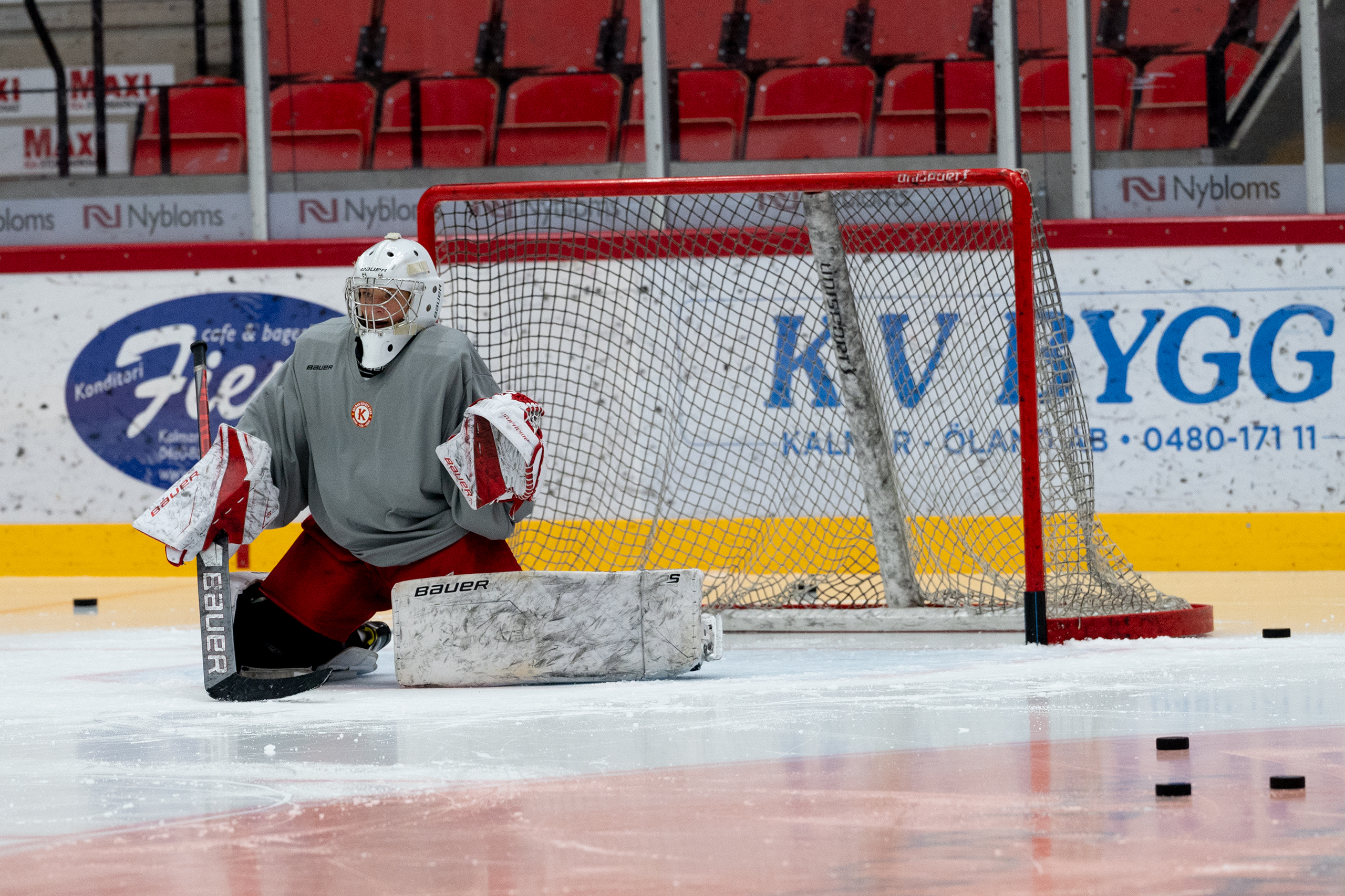 Ishockeymålvakt sittandes framför ishockeymål till vänster i bild. På isen ligger puckar.