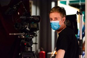 En man som sitter bakom en kamera och har munskydd på sig.
