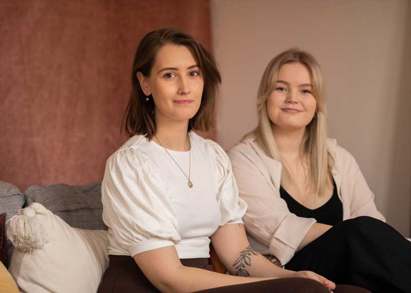 Tess och Linda sitter ner framför en rosa bakgrund.