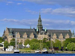 En stor byggnad med ett torn i mitten. Träd framför och vatten med en båt.