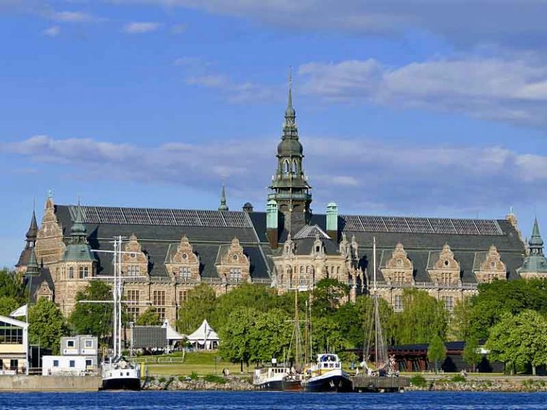 En stor byggnad med ett torn i mitten. Träd framför och vatten med en båt.