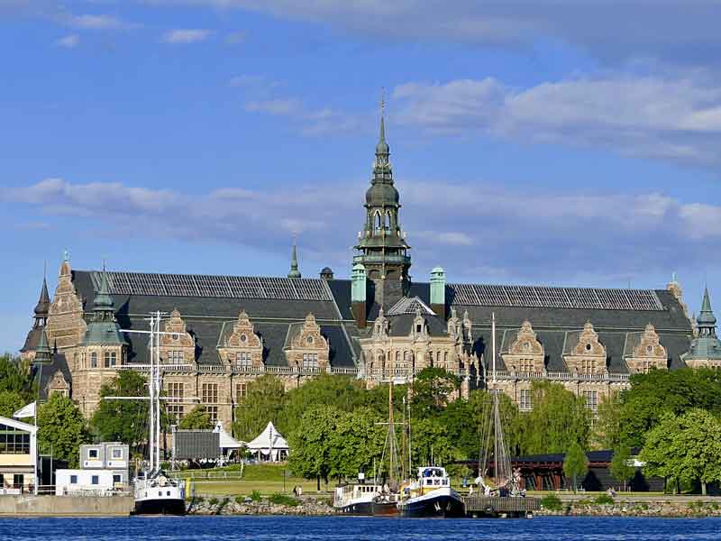 En stor byggnad med ett torn i mitten. Träd framför och vatten med en båt.