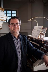 En man står bredvid en orgel. Han har rutig skjorta, mörkblå kavaj och glasögon.