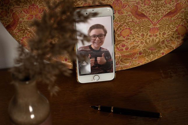 En kille på en mobilskärm som ler och håller upp tummarna. En blomma i förgrunden och en gul bricka med mönster bakom.